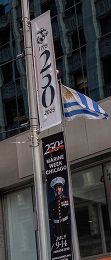 A street pole displays Marine Week Chicago banners and a blue-and-white flag. The banners mention the U.S. Marine Corps’ 250th anniversary in 2025 and promote Marine Week Chicago, July 9–14.