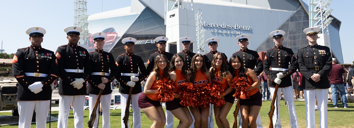 A group of U.S. Marines in dress uniforms stands in a row behind five cheerleaders in maroon and orange outfits. They pose outside the Mercedes-Benz Stadium on a sunny day.