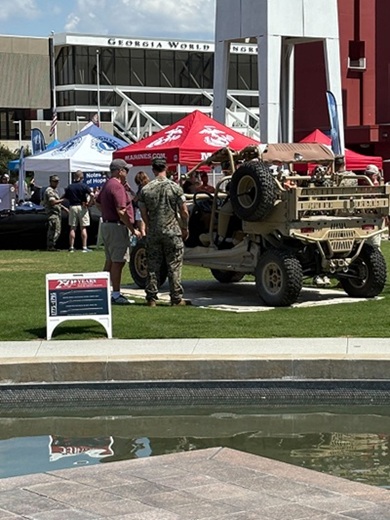 A military vehicle is displayed on grass near a fountain, with people standing around. Red, white, and blue tents labeled "Marines.com" and "Navy" are set up in the background near a large building.