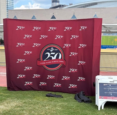 A maroon banner displays “250 United States Marine Corps” logos, set up outdoors on grass with a large building in the background. A small white stand and a rolled banner are on the ground nearby.