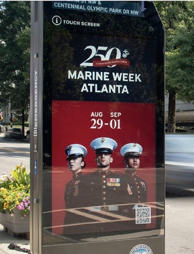 A street kiosk displays a Marine Week Atlanta poster featuring three U.S. Marines in dress uniforms, event dates August 29 to September 1, and the Marine Corps 250th anniversary logo. Trees and city street are in the background.